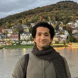 Ein Mann mit lockigem Haar steht an einem Flussufer. Er trägt eine graue Jacke und einen Schal. Im Hintergrund sind Häuser an einem Hang und herbstliche Bäume sichtbar.