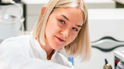 Eine junge Frau mit blonden, schulterlangen Haaren schaut direkt in die Kamera. Sie trägt einen weißen Laborkittel und arbeitet offensichtlich in einem wissenschaftlichen Umfeld, umgeben von Gerätschaften und Laborutensilien. Ihr Ausdruck ist freundlich und engagiert.
