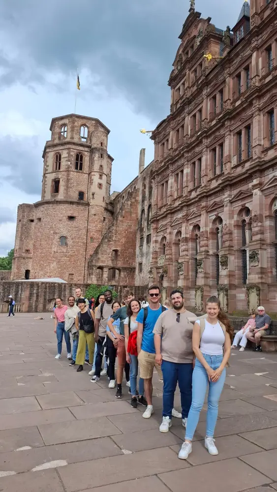 Eine Gruppe von zehn Personen steht in einer langen Reihe vor einer historischen Burg. Im Hintergrund sind die beeindruckende Architektur der Burg und ein bewölkter Himmel zu sehen. Einige Menschen sitzen entspannt auf einer Bank in der Umgebung.
