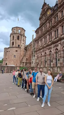 Eine Gruppe von zehn Personen steht in einer langen Reihe vor einer historischen Burg. Im Hintergrund sind die beeindruckende Architektur der Burg und ein bewölkter Himmel zu sehen. Einige Menschen sitzen entspannt auf einer Bank in der Umgebung.