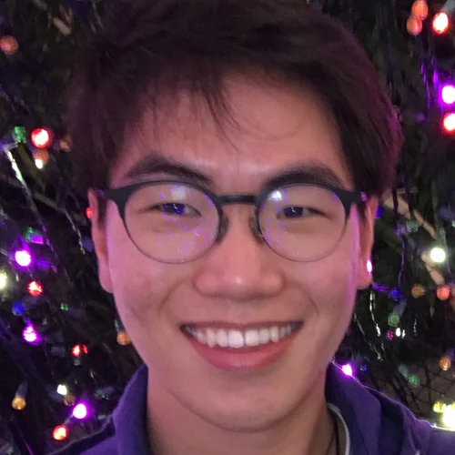 A young person with short dark hair and glasses smiles warmly at the camera. Behind them, soft, colorful lights are softly glowing, creating a festive atmosphere.