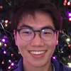 A young person with short dark hair and glasses smiles warmly at the camera. Behind them, soft, colorful lights are softly glowing, creating a festive atmosphere.