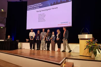 Eine Gruppe von sechs Personen auf einer Bühne bei einer Preisverleihung. Im Hintergrund steht ein Präsentationsbildschirm mit dem Titel "Young Investigator Forum". Die Teilnehmer halten Medaillen oder Urkunden in den Händen, während ein Moderator neben ihnen steht.