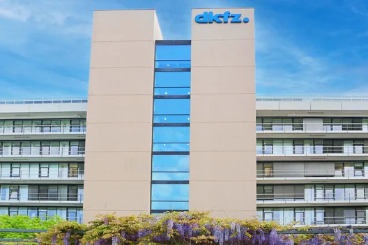 A modern, multi-story building features large glass windows and a prominent sign displaying the name "dkfz." The structure is surrounded by greenery and flowering plants, emphasizing an inviting environment. The sky is clear and blue, enhancing the overall appearance of the facility.