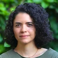 Eine Frau mit lockigem, dunklem Haar schaut direkt in die Kamera. Sie trägt ein schlichtes, grünes Oberteil und hat einen neutralen Ausdruck. Im Hintergrund sind unscharfe grüne Pflanzen zu sehen.