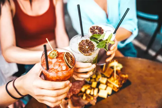 Drei bunte Cocktails werden über einer Snackplatte angestoßen.